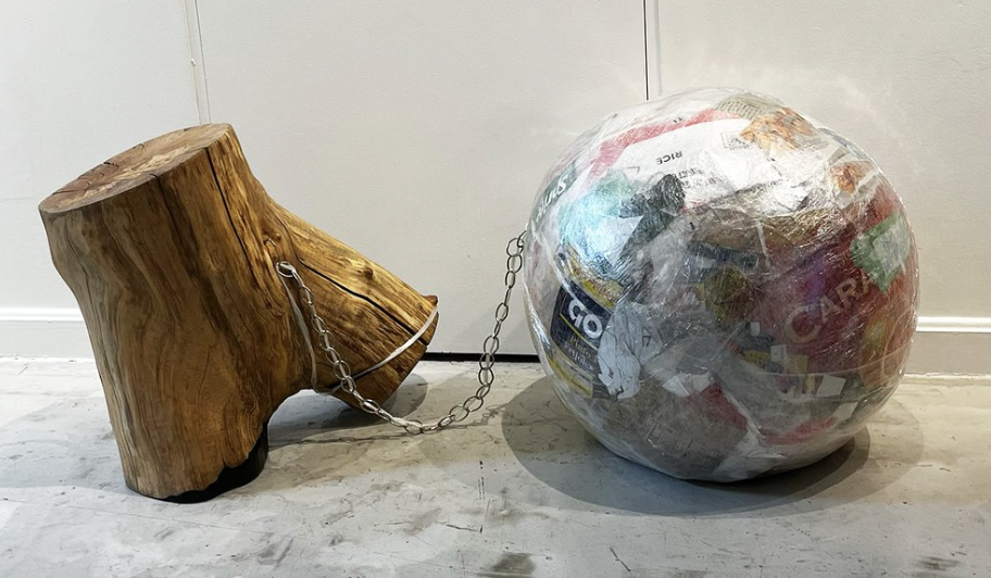 A globe crafted from discarded plastic found on a reservation beach. The trunk, resembling a torso, from a fallen tree amidst the aftermath of a recent storm. A chain tethers the sculpture and globe, symbolizing the interconnectedness between humanity and waste.